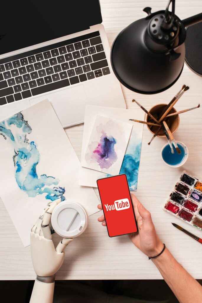 cropped view of designer at office desk with robot hand holding smartphone with youtube app on